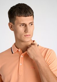 Polo di colore arancione chiaro realizzato in tessuto testurizzato, con colletto a righe bianche e nere e logo ricamato sopra il lato sinistro del petto.