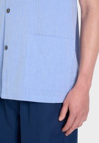 Camicia a righe blu chiaro realizzata in un tessuto morbido, con una chiusura a bottoni sul davanti, maniche corte e una tasca sul petto. Abbinata a pantaloni corti blu scuro.