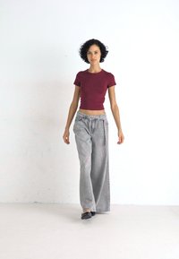 Bordo izrezan T-majica v kombinaciji s svetlo sivo širokimi hlačami. Mehak material z rahlo teksturo. Model nosi črne ravne čevlje, postavljen proti beli podlagi.