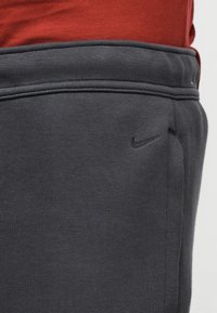 Pantaloni de trening gri închis cu o textură netedă, având un mic logo negru Nike și o talie cu elastic striat. Bluza roșie este parțial vizibilă deasupra.