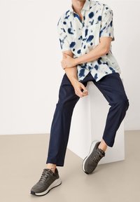 Kortärmad skjorta med knappar i lätt tyg, prydd med blå och marinblå färg-liknande mönster, kombinerad med mörka byxor och grå sneakers.