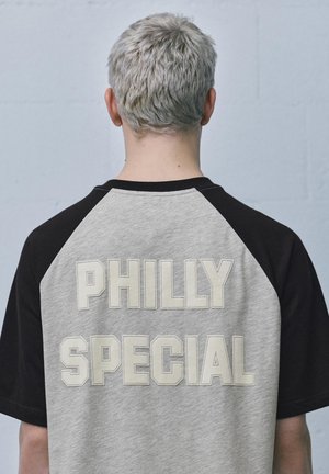 Oseba s kratkimi svetlo sivimi lasmi nosi sivo-črno majico z velikimi črkami »PHILLY SPECIAL« na hrbtu.