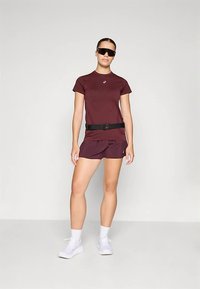 Nainen, jolla on päällä maroon-värinen urheilupaita ja shortsit, valkoiset lenkkarit, musta vyö ja suuret mustat urheililasit, seisoo yksinkertaista taustaa vasten.