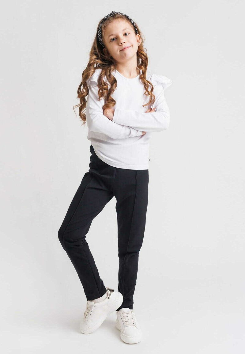 Biała bluzka z długim rękawem i falbankami na ramionach, zestawiona z czarnymi dopasowanymi spodniami i białymi trampkami. Prosty design bez wzorów.