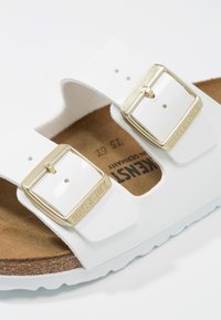 Sandalias Birkenstock blancas con dos correas ajustables y hebillas doradas, una plantilla de corcho y una suela blanca texturizada.