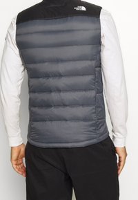 Gepolsterte graue Weste mit schwarzer Schulterpartie, mit horizontalem Steppmuster und Logo auf der Rückseite, kombiniert mit einem langärmligen Hemd.