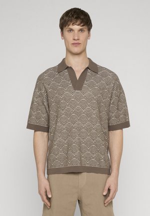 Hombre con camisa tipo polo de manga corta de punto con patrón marrón y pantalones beige, de pie frente a un fondo claro y liso mirando hacia adelante.