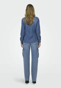 Frau mit langen blonden Haaren, die ein Jeanshemd und eine hellblaue Cargo-Jeans trägt, steht mit dem Rücken zur Kamera vor einem einfarbigen Hintergrund.