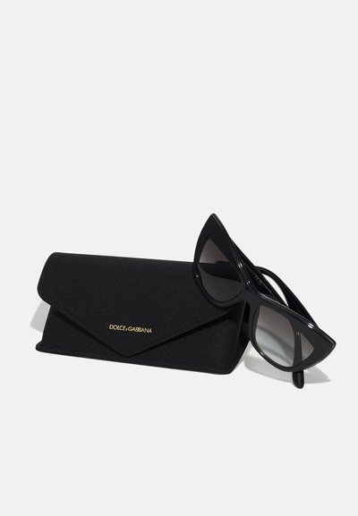 Black sunglasses with sharp, angular frames next to a black fabric case featuring a gold logo, both showcasing a sleek, minimalist design.