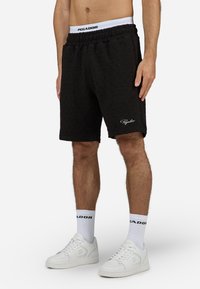 Shorts matelassés noirs avec une taille élastique et le nom "PAGADOR" brodé en blanc. Portés avec des baskets blanches et des chaussettes.