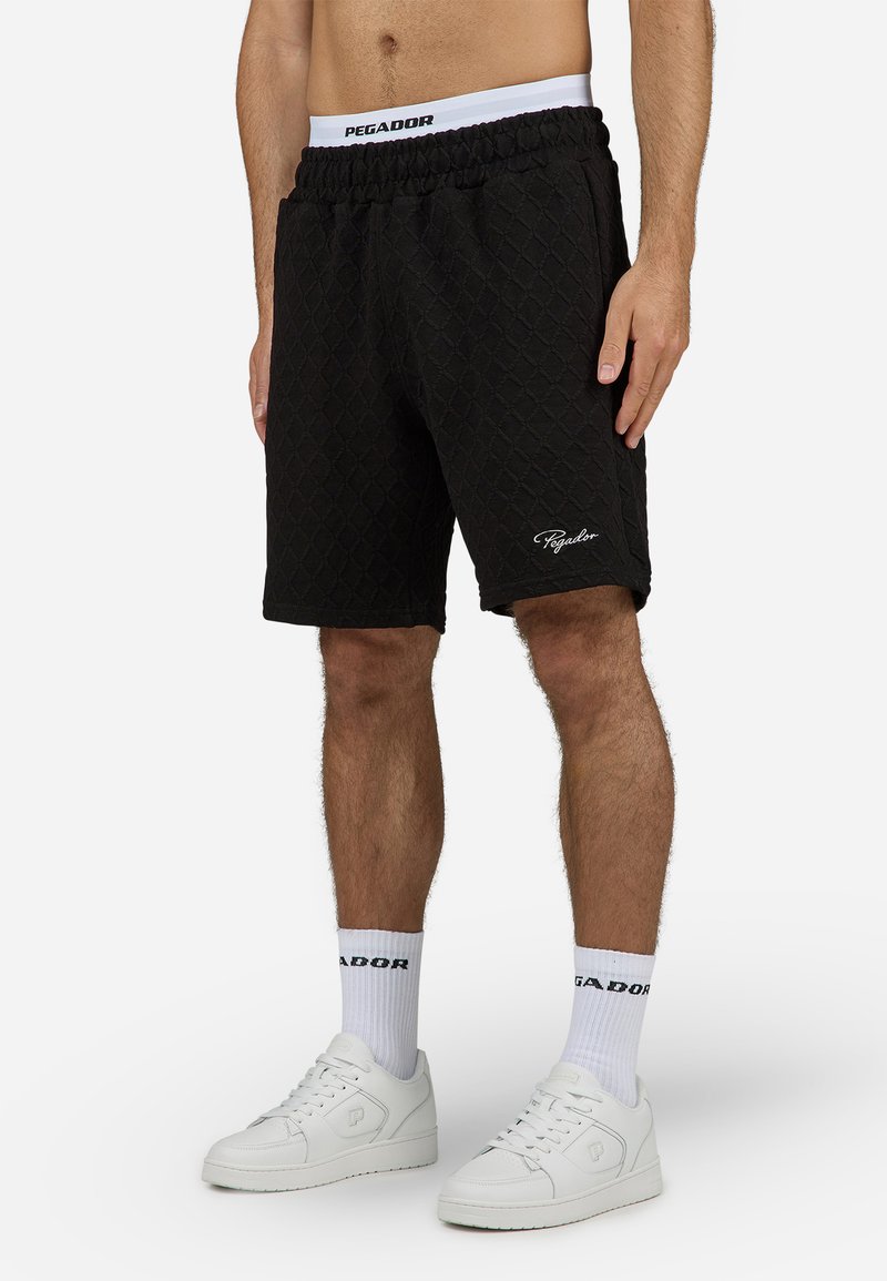 Shorts matelassés noirs avec une taille élastique et le nom "PAGADOR" brodé en blanc. Portés avec des baskets blanches et des chaussettes.