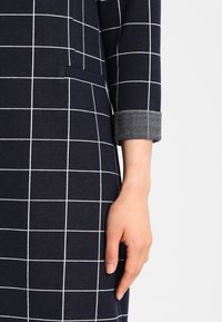 Robe bleu marine à motif quadrillé blanc, avec un revers plié, une poche latérale et une texture lisse. Main visible près de l'ourlet.