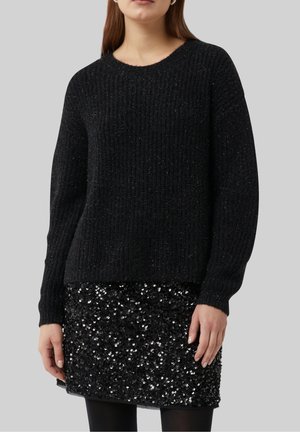Femme portant un pull en maille côtelée noire associé à une mini-jupe noire à sequins et des collants noirs, sur un fond gris uni.