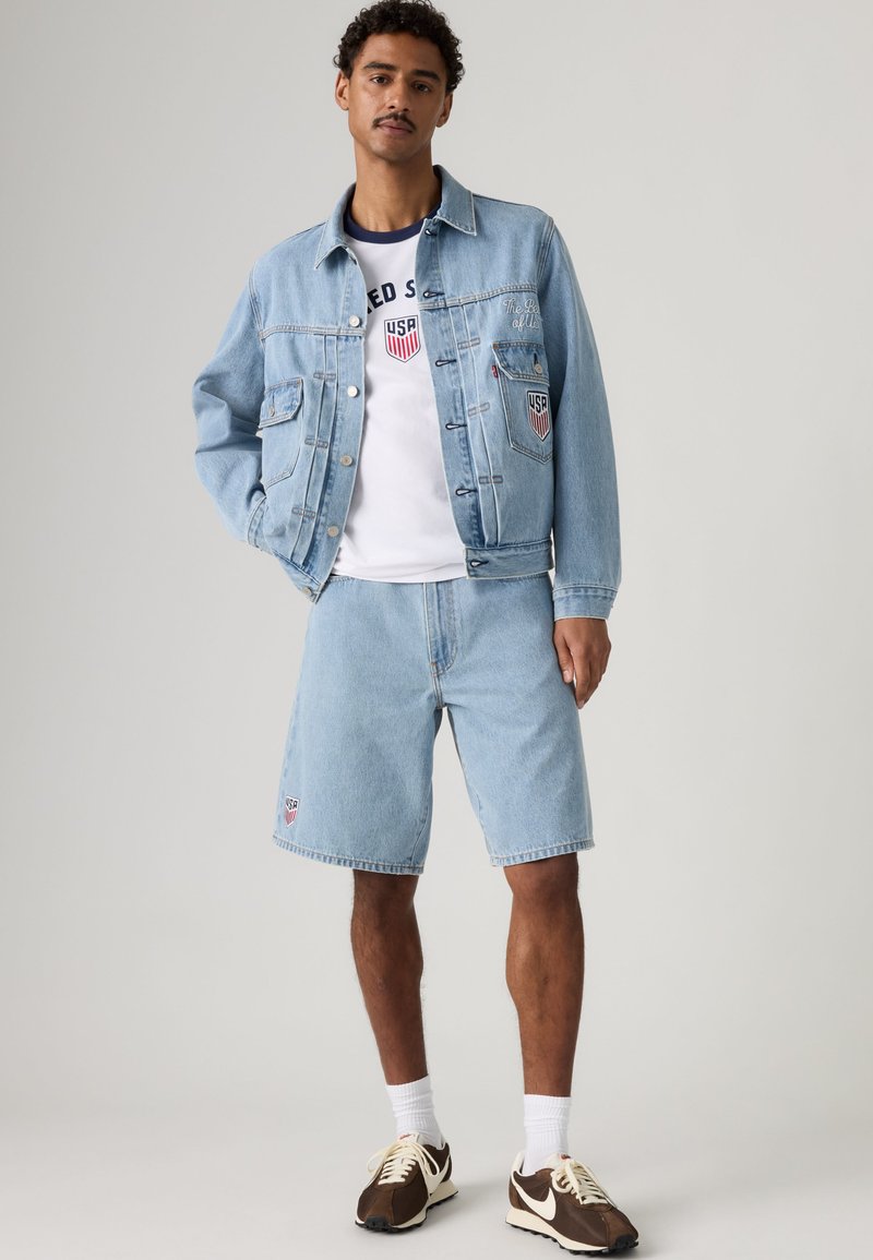 Mann trägt hellblaue Jeansjacke und Shorts mit weißem USA-Emblem, weißes Grafik-T-Shirt, weiße Socken und braune Sneaker, steht mit der Hand in der Tasche.