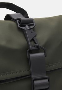 Détail d'un sac étanche vert avec une surface mate, comportant un clasp en plastique noir, une sangle et une couture contrastante le long du bord.