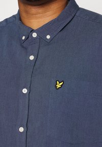 Granatowa koszula na guziki z teksturowanej tkaniny, z naszytą kieszenią i żółtym haftowanym logo na piersi. Guziki są białe.