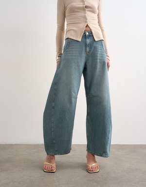 Személy világos bézs színű, gombos felsőt, magas derekú, bőszárú kék farmert és sárga pántos szandált visel, szürke padló és fehér fal háttér előtt.