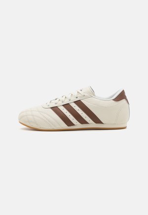 White leather sneaker with brown stripes, featuring a rounded toe, perforated detailing, and a gum rubber sole.