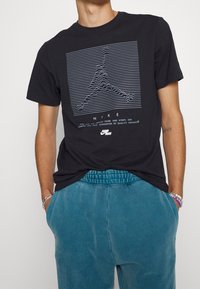 Černé bavlněné tričko s šedým grafickým designem a vlnitými liniemi, s nápisem "NIKE" umístěným na přední straně. V kombinaci s modrými tepláky.