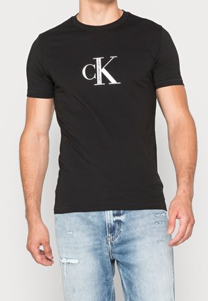 Schwarzes Baumwoll-T-Shirt mit einem weißen "CK"-Logo auf der Brust. Kurzarm, runder Halsausschnitt und schlanker Schnitt. Getragen mit ausgewaschenen blauen Jeans.
