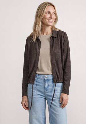 Kvinde med langt blond hår iført en mørk knappet cardigan over en beige top og lyseblå jeans, smilende og kiggende mod højre.