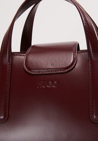 Bordeauxrode leren handtas met een gladde textuur, voorzien van een klepsluiting en een geperst "HUGO"-logo. Korte, afgeronde handvatten.