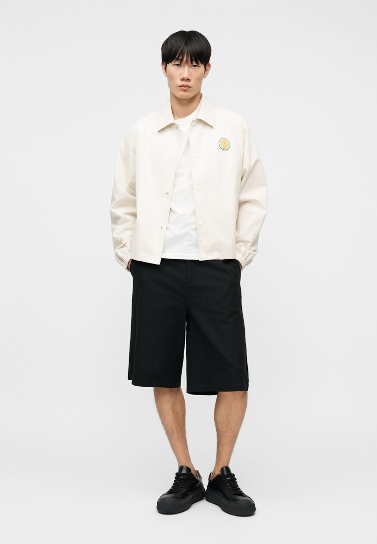 Jeune homme debout portant une veste blanche à boutons avec un logo, un t-shirt blanc, un short noir au genou et des baskets noires sur un fond blanc.