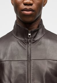 Κοντινό πλάνο ενός ατόμου που φοράει σκούρο καφέ δερμάτινο μπουφάν με φερμουάρ στον ψηλό γιακά, δείχνοντας το κάτω μέρος του προσώπου και τον λαιμό.