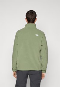 Muškarac nosi maslinasto zeleni flis s jaknom s logom The North Face na gornjem dijelu leđa, okrenut leđima, stoji uz jednostavnu svijetlu pozadinu.