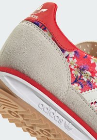 Adidas-sneaker med en grå mockatopp, livfull blommönster i rött och lila, vit tre-stripes design, gummisula.
