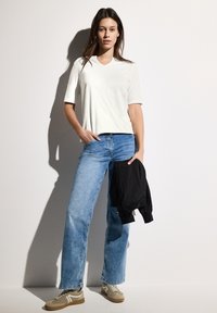 Haut blanc à manches courtes, coupe décontractée, associé à un jean large bleu clair. Baskets beige avec accents blancs. Veste noire tenue à la main.