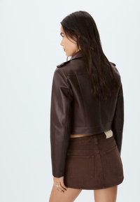 Donna con lunghi capelli scuri, che indossa un giubbotto di pelle marrone scuro corto e una gonna di jeans marrone coordinata, in piedi di lato contro uno sfondo semplice.