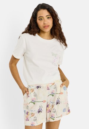 Mujer con cabello largo y rizado que lleva una camiseta blanca con un pequeño gráfico de mariposa y pantalones cortos con estampado floral, manos en los bolsillos.