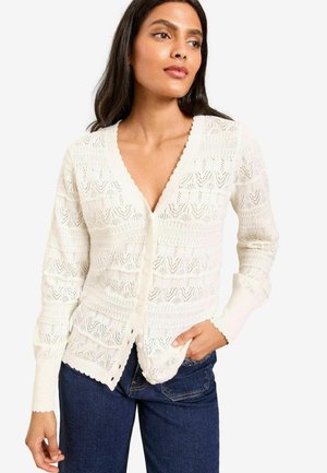 Cream, knitted cardigan featuring a deep V-neck, scalloped edges, and intricate openwork patterns. Buttons down the front, fitted sleeves.