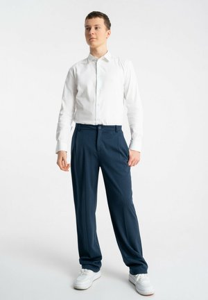 Młoda osoba stojąca, ubranа w białą koszulę na guziki, granatowe spodnie i białe trampki, na tle gładkiej białej ściany.