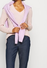 Suéter rosa claro con escote en V, que presenta un acabado texturizado. Alrededor de los hombros lleva una bufanda lila. Combinado con pantalones azul oscuro.