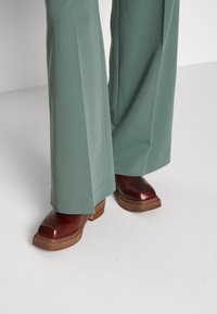 Pantalons larges verts en tissu lisse, assortis à des sabots à talons épais bruns avec une finition brillante et des semelles en bois.