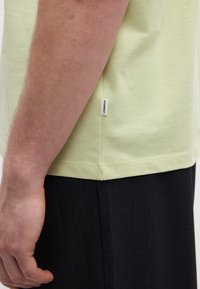 Αριστερό χέρι ενός ατόμου που φοράει ανοιχτοκίτρινο πουκάμισο με ετικέτα "LANDEBERGH" και σκούρο παντελόνι, εμφανιζόμενο από το πλάι με φόντο λευκό.