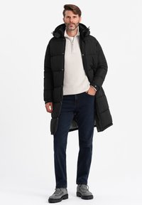 Sort isoleret dunjakke med hætte, sidelommer og trykknaplukning, båret over en lys cremefarvet sweater og mørke jeans, suppleret med grå støvler.