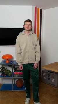 Ung mann i beige hettegenser med glidelås og grønne treningsbukser står innendørs nær TV, oransje lampe, basketball og koffert med klistremerker.