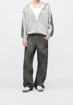 Personne portant une veste à capuche grise zippée avec des bandes noires sur les manches, une chemise blanche, un pantalon noir à motifs et des baskets blanches, debout les mains dans les poches.