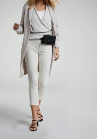 Un long cardigan gris clair superposé à un haut gris clair, associé à un jean blanc cropped et des talons aiguilles noirs à lanières. Sac à main noir accroché.