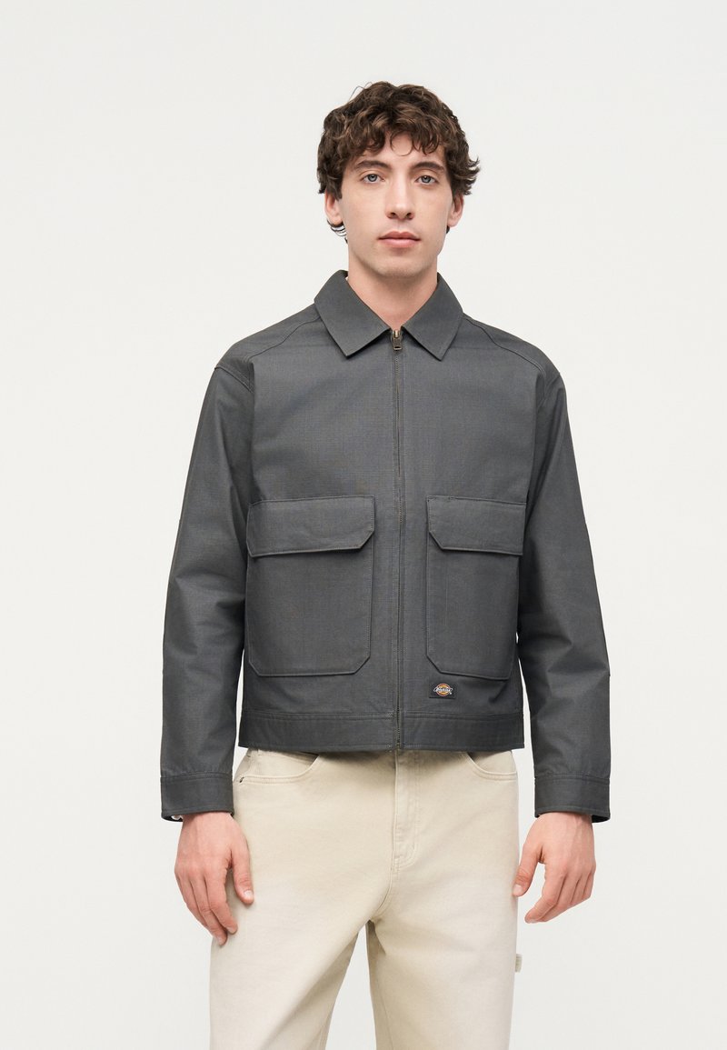 Junger Mann trägt eine dunkelgraue Jacke mit Reißverschluss und zwei vorderen Klappentaschen sowie helle beige Hosen, steht vor einem schlichten hellen Hintergrund.