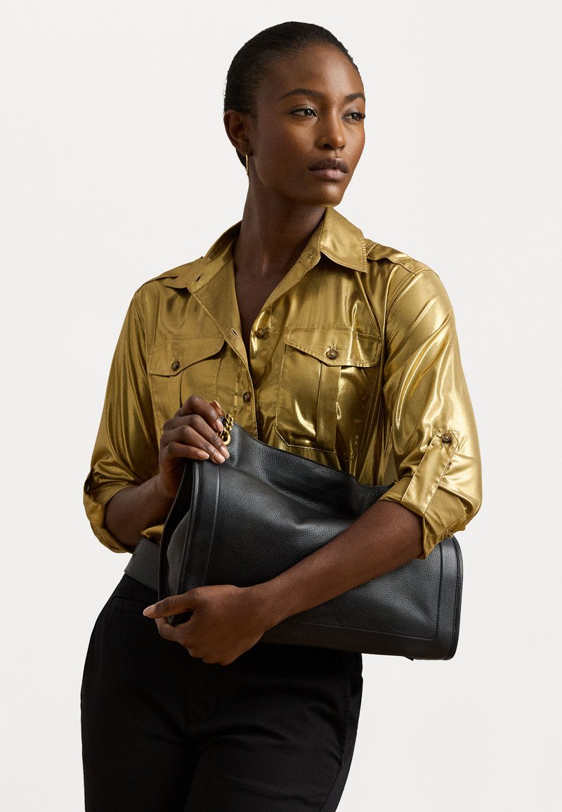 Melnas ādas soma ar vaļīgu dizainu, kas izceļas ar gludu tekstūru un maigiem šuvumiem. Modelis to tur pie zelta satīna krekla.