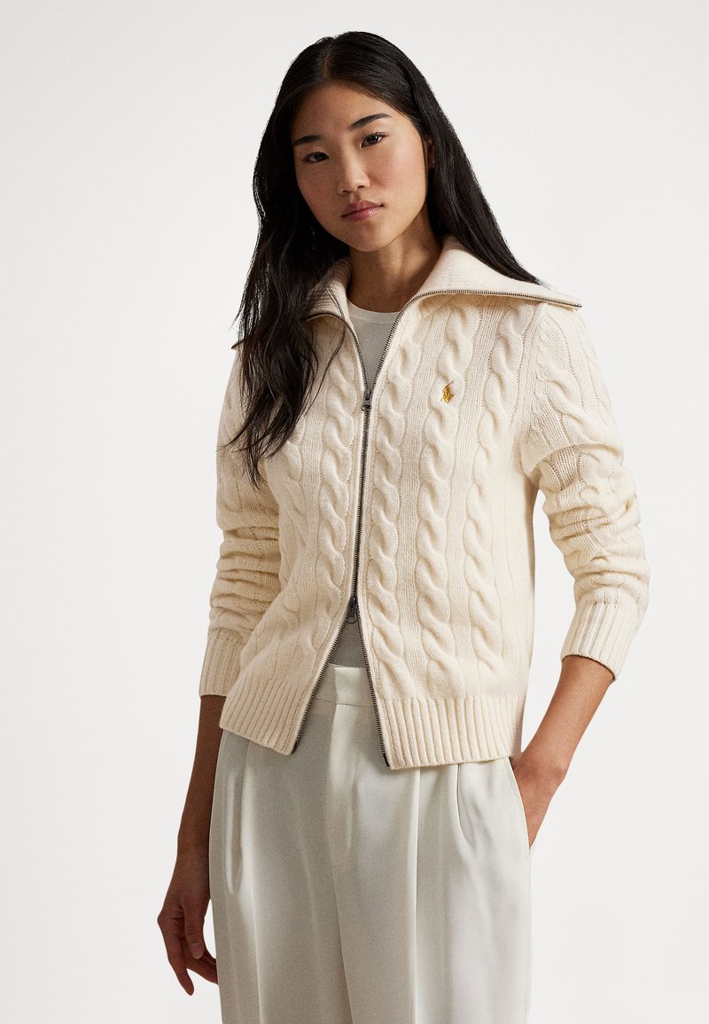 Pulover v kremo barvi s pletenim vzorcem kabel, s polovičnim zadrgo na ovratniku, rebrastimi manšetami in spodnjim robom, na levem prsnem delu z majhnim logotipom.