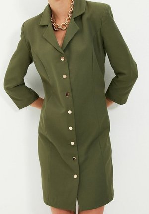 Vrouw die een olijfgroene jurk met knopen draagt, met driekwartmouwen en een stevige gouden ketting.