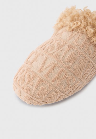 Chaussure beige à enfiler avec un design en relief et en lettres texturées. Accent duveteux brun clair en haut. Bout rond et matériau en tissu doux.