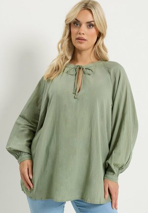 Vaaleahiuksinen nainen, jolla on yllään löysä, pitkähihaisen salviavihreä pusero, jossa on solmittava kaulanauha, ja vaaleansiniset farkut, seisomassa yksiväristä taustaa vasten.