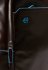 Bolso de cuero marrón con un cierre metálico, ribetes azules en los bolsillos y un logo azul. Textura suave con costuras detalladas.