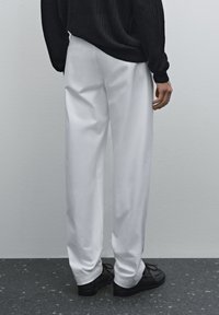Hvide, vide bukser lavet af glat stof, parret med sorte sko, set fra ryggen. Baggrunden er neutral og minimalistisk.